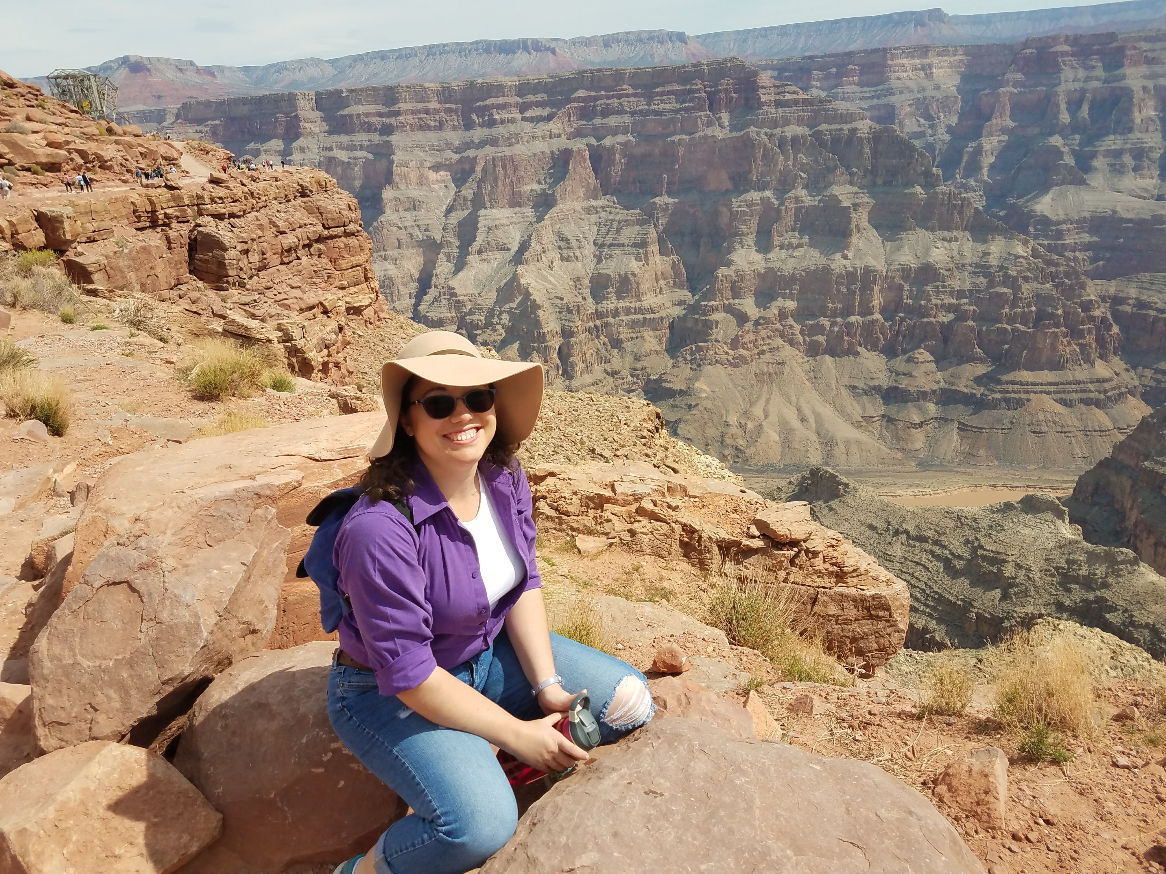 Woman in purple shirt and a sun hat sits at the edge of the Grand Canyon